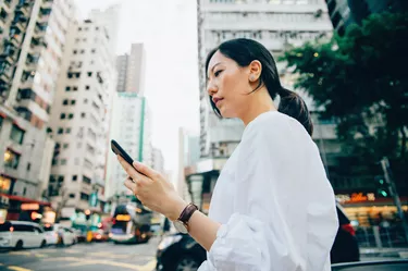 Confidence young Asian woman using mobile phone while crossing road in downtown city, against busy city traffic and urban city buildings