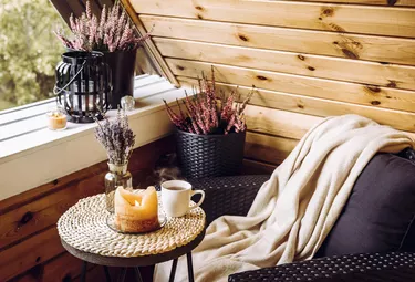 Cute autumn home decor arrangement. Tiny wooden cabin balcony with heather flowers in pot, lavender in bottle vase, candlelight flame, soft beige plaid waiting on comfortable garden furniture chair.