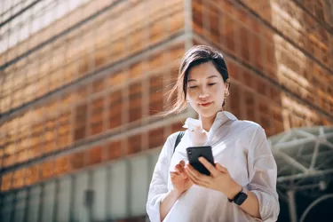 Low angle portrait of young Asian businesswoman checking emails on smartphone outside office building in financial district. With contemporary corporate skyscrapers in background. Making business connections throughout the city