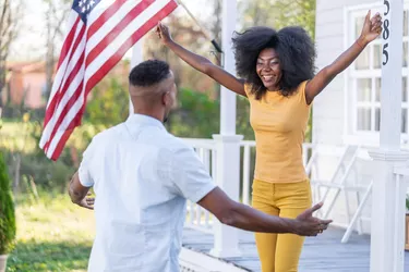 Smiling afro couple with wide open arms for big hug in front their new house with the american flag on the porch